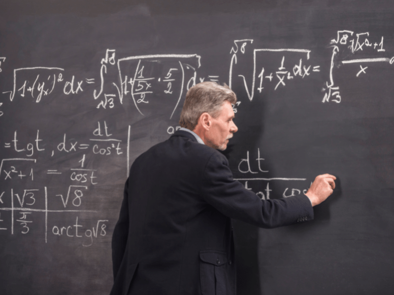 A professor standing in front of a black board with chalk in hand.
